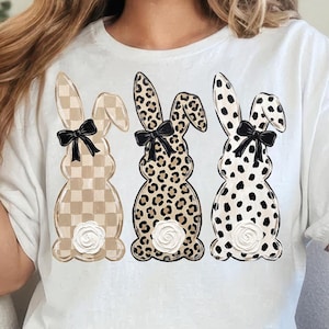 May include: White t-shirt featuring three bunny designs with different patterns: checkered, leopard print, and polka dots. Each bunny has a black bow and a floral accent. The shirt is worn with blue jeans.