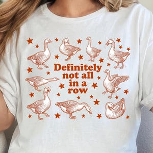 May include: White t-shirt with a rust-colored goose and star design. The text "Definitely not all in a row" is printed in the center. The shirt is a casual, comfortable style.