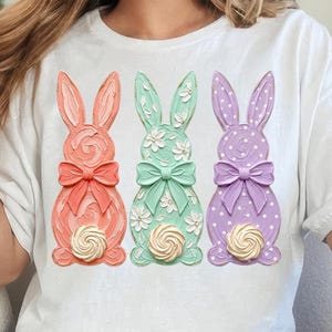 May include: White t-shirt featuring three pastel-colored Easter bunny designs. Each bunny has a bow and a swirl detail at the bottom. The bunnies are in shades of coral, mint green, and lavender, with the third bunny having a polka dot pattern.