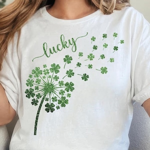 Könnte beinhalten: Weißes T-Shirt mit einem grünen Löwenzahnmotiv, das aus Kleeblättern besteht, mit dem Wort "lucky" in Schreibschrift. Einige Kleeblätter werden weggeblasen dargestellt. Ein lässiges Oberteil, passend für St. Patrick's Day oder den täglichen Gebrauch.