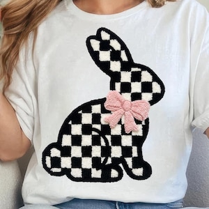 Könnte beinhalten: Weißes T-Shirt mit einem schwarz-weiß karierten Hasenmuster und einer rosa Schleife. Die Hasengrafik befindet sich auf der Vorderseite des Shirts. Das Shirt hat kurze Ärmel und einen Rundhalsausschnitt.