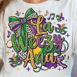 May include: White t-shirt with a vibrant graphic design. The design features the text "La Weezy Ana" in green and gold, complemented by a bow, musical notes, and Mardi Gras mask motifs. A casual wear item.