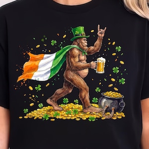 May include: Black t-shirt featuring a Bigfoot character wearing a green hat and Irish flag cape, holding a beer mug. The design includes a pot of gold, shamrocks, and gold coins, celebrating St. Patrick's Day.