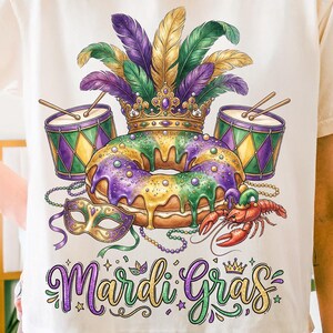 Könnte beinhalten: Cremefarbenes T-Shirt mit einem farbenfrohen Mardi-Gras-Design. Die Grafik zeigt einen King Cake, eine Krone, Federn, Trommeln, eine Maske und einen Krebs. Die Worte "Mardi Gras" sind in einer dekorativen Schrift geschrieben.