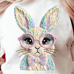 May include: White sweatshirt featuring a colorful, embroidered bunny wearing pink glasses and a bow tie. The bunny has long eyelashes and a detailed design with pastel colors, perfect for Easter or spring.