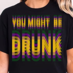 May include: Black t-shirt featuring the text "YOU MIGHT BE DRUNK" in layered yellow, purple, and green lettering. The top has short sleeves and a crew neck.