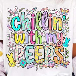 Może przedstawiać: Biała koszulka z napisem "Chillin' with my Peeps" w kolorowych literach. Projekt zawiera grafikę w tematyce wielkanocnej, taką jak króliki, jajka, motyle i symbole medyczne. Swobodny, świąteczny element garderoby.