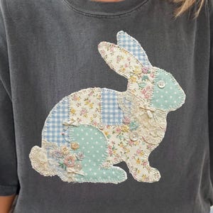 Peut inclure: T-shirt gris foncé avec un motif de lapin en patchwork. Le lapin est fait de tissu floral, vichy et à pois avec des détails en dentelle et des boutons. Le t-shirt a des manches courtes et un col rond.