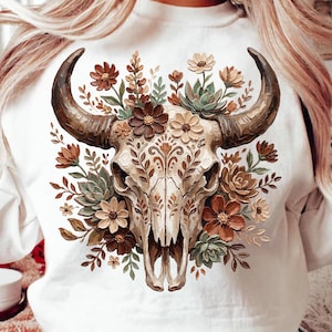 May include: White sweatshirt featuring a detailed graphic of a bull skull adorned with flowers and foliage in shades of brown, tan, and green. The skull has large, curved horns. The design is centered on the front of the shirt.