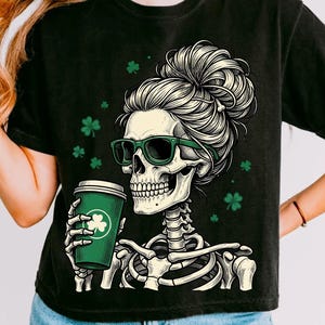 Op de afbeelding: Zwart T-shirt met een skeletontwerp met een groene zonnebril en een knot, met een groene koffiebeker met een klavertje. Groene klavers zijn verspreid. Een casual T-shirt.