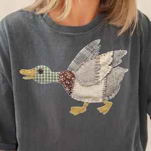Könnte beinhalten: Dunkelgraues T-Shirt mit einem Enten-Design im Patchwork-Stil. Die Ente hat einen grün-weiß karierten Kopf, einen braun-weiß gemusterten Körper und grau-weiße Flügel. Die Füße der Ente sind gelb.