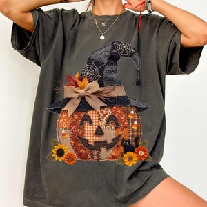 May include: A charcoal gray short-sleeve t-shirt featuring a graphic of a patchwork pumpkin wearing a witch hat. The design includes a tan bow, autumn leaves, sunflowers, candy corn, a black cat, and a spider hanging from the hat brim.