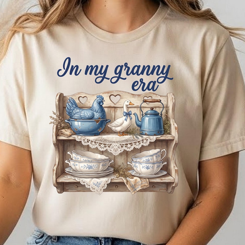Może przedstawiać: Beżowa koszulka z napisem "In my granny era" w kolorze granatowym. Grafika przedstawia drewnianą p&oacute;łkę z niebieską kurą, białą kaczką z niebieską kokardą, niebieskim czajnikiem i filiżankami.