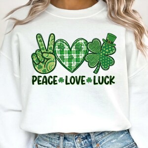 May include: White sweatshirt with a green graphic design featuring a peace sign, heart, and shamrock. The words "PEACE LOVE LUCK" are printed in green below the symbols. The design incorporates various green patterns and a small green hat.