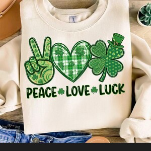 May include: Cream-colored sweatshirt with a St. Patrick's Day graphic. The design includes a peace sign, a heart, and a shamrock, all in shades of green. The words "PEACE LOVE LUCK" are printed below the symbols.