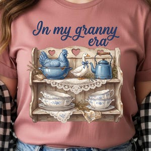 Może przedstawiać: R&oacute;żowa koszulka z napisem "In my granny era". Koszulka przedstawia dekoracyjną p&oacute;łkę z niebieską kurą, białą kaczką z niebieską kokardą, niebieskim imbrykiem i filiżankami. P&oacute;łka ma styl vintage.
