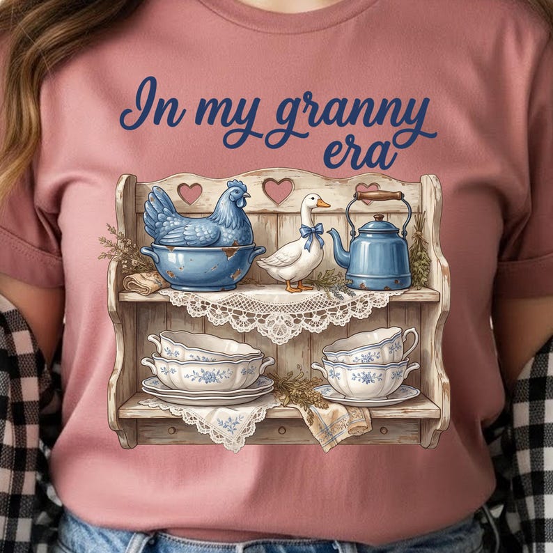 Może przedstawiać: R&oacute;żowa koszulka z napisem "In my granny era". Koszulka przedstawia dekoracyjną p&oacute;łkę z niebieską kurą, białą kaczką z niebieską kokardą, niebieskim imbrykiem i filiżankami. P&oacute;łka ma styl vintage.