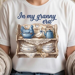 Może przedstawiać: Biała koszulka z granatowym napisem "In my granny era". Grafika przedstawia drewnianą p&oacute;łkę z niebiesko-białymi filiżankami, niebieską kurą, białą kaczką i niebieskim imbrykiem.