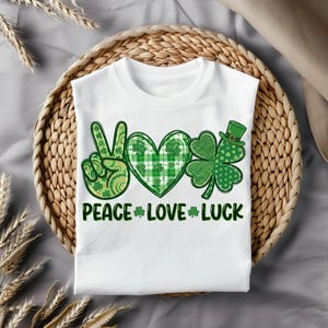 May include: White t-shirt featuring a green peace sign, heart, and shamrock design. The heart has a plaid pattern, and the shamrock has a leprechaun hat. The words "PEACE LOVE LUCK" are printed below the graphics in green.