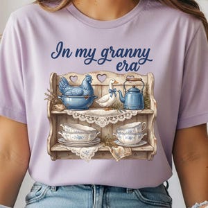Może przedstawiać: Jasnofioletowa koszulka z napisem "In my granny era". Grafika przedstawia drewnianą p&oacute;łkę z niebieskim kurczakiem w misce, białą kaczką, niebieskim imbrykiem i filiżankami do herbaty z kwiatowymi wzorami.