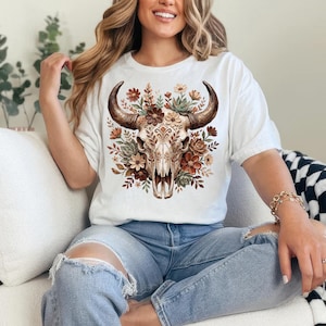 May include: White t-shirt featuring a detailed watercolor illustration of a bull skull adorned with flowers in shades of brown, orange, and green. The skull has large, curved horns. The shirt is a casual style.
