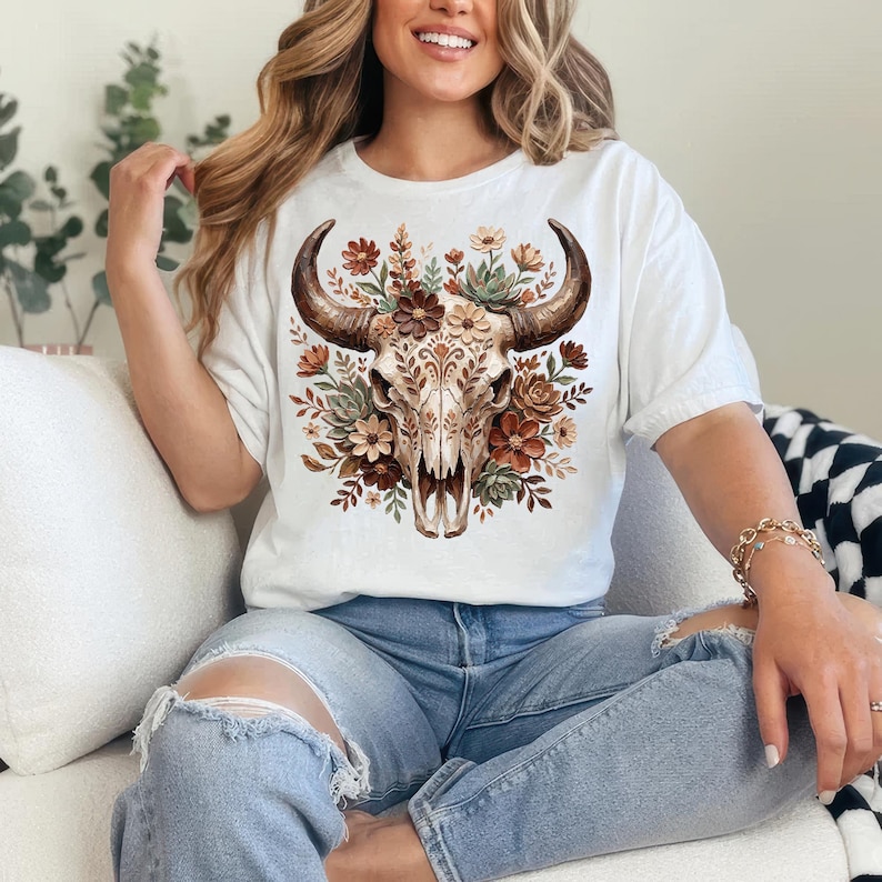 May include: White t-shirt featuring a detailed watercolor illustration of a bull skull adorned with flowers in shades of brown, orange, and green. The skull has large, curved horns. The shirt is a casual style.