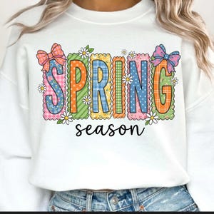 May include: White sweatshirt with the word "SPRING" in colorful, patterned letters. The letters are outlined with scalloped edges and decorated with daisies and bows. The word "season" is written in a cursive font below.
