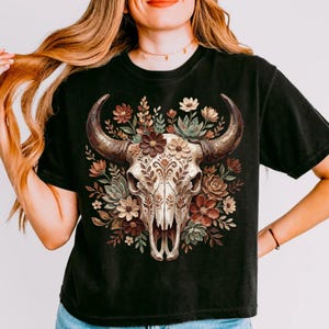 May include: Black t-shirt featuring a detailed graphic of a cow skull adorned with flowers and foliage in shades of brown, cream, and green. The skull has large, curved horns. The design is centered on the shirt.