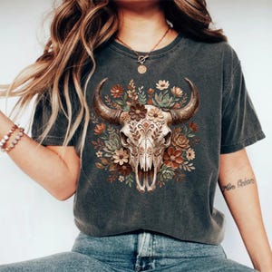 May include: Dark gray t-shirt featuring a detailed illustration of a bull skull adorned with floral elements in shades of brown, beige, and green. The bull skull has intricate patterns. The shirt is a casual wear item.