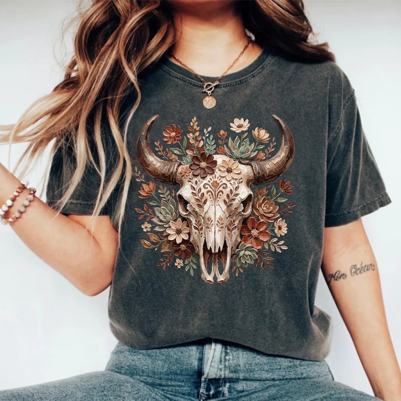 May include: Dark gray t-shirt featuring a detailed illustration of a bull skull adorned with floral elements in shades of brown, beige, and green. The bull skull has intricate patterns. The shirt is a casual wear item.