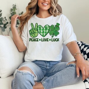 May include: White t-shirt with a St. Patrick's Day graphic. The design includes a peace sign, a heart, and a shamrock, all in shades of green. The words "PEACE LOVE LUCK" are printed below the symbols.
