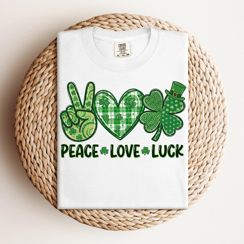 May include: White t-shirt featuring a green peace sign, heart, and shamrock design. The heart has a plaid pattern. The words "PEACE LOVE LUCK" are printed in green below the graphics. The shirt is folded on a woven basket.