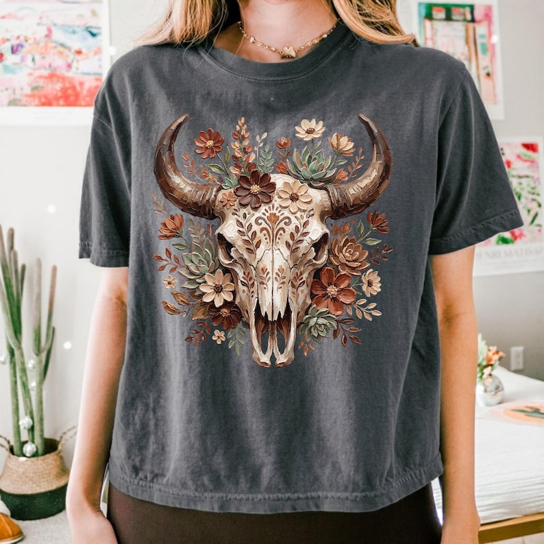 May include: Dark gray t-shirt featuring a detailed illustration of a cow skull adorned with flowers. The skull is cream-colored with brown horns, surrounded by burgundy, cream, and green floral elements. The shirt has short sleeves and a crew neck.