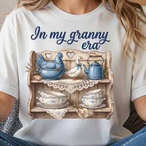 Może przedstawiać: Biała koszulka z napisem "In my granny era" nad ilustracją p&oacute;łki. P&oacute;łka przedstawia niebieskiego kurczaka w misce, białą kaczkę z niebieską kokardą, niebieski czajnik i filiżanki.