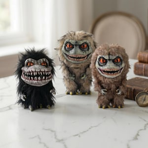 May include: Three Critters-inspired figurines with menacing expressions. The figurines have sharp teeth, red eyes, and shaggy fur in black, tan, and brown. They stand on a white surface, evoking a horror or fantasy theme.