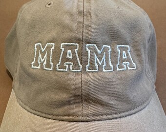 May include: A tan baseball cap with the word "MAMA" embroidered in white block letters. The hat has a curved brim and a fabric-covered button on top. The hat is made of a durable, cotton-like material.