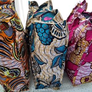 Könnte beinhalten: Mehrere Stoff-Tragetaschen mit lebendigen, afrikanisch inspirierten Mustern. Die Taschen weisen kräftige Farben wie Orange, Türkis und Pink mit geometrischen und floralen Designs auf. Die Taschen stehen aufrecht und zeigen ihre Form und die komplizierten Muster.