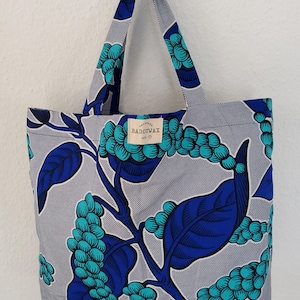 Könnte beinhalten: Eine Tragetasche mit einem sich wiederholenden botanischen Muster aus blauen und türkisfarbenen Beeren und Blättern auf hellgrauem Grund mit feinen diagonalen Linien. Ein rechteckiges Stoffetikett trägt den Text BADOUWAX und ein Herz.