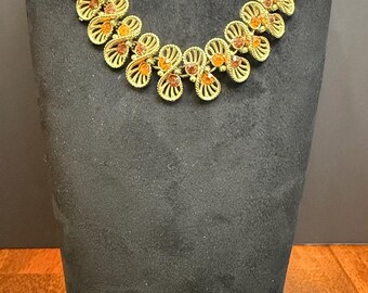 Peut inclure: Ensemble collier et broche dorés. Le collier présente une série de maillons ajourés en forme de fleurs avec des accents de couleur ambre. La broche assortie a un design similaire. Les bijoux sont exposés sur un mannequin noir.