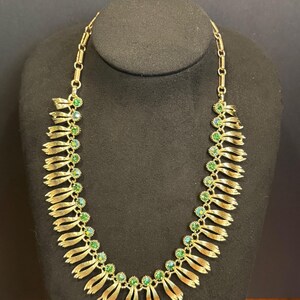 May include: Gold-toned necklace with a series of layered, ribbon-like elements. Each ribbon section is adorned with a cluster of green and teal-colored gemstones. The necklace is displayed on a black mannequin, showcasing its design and details.