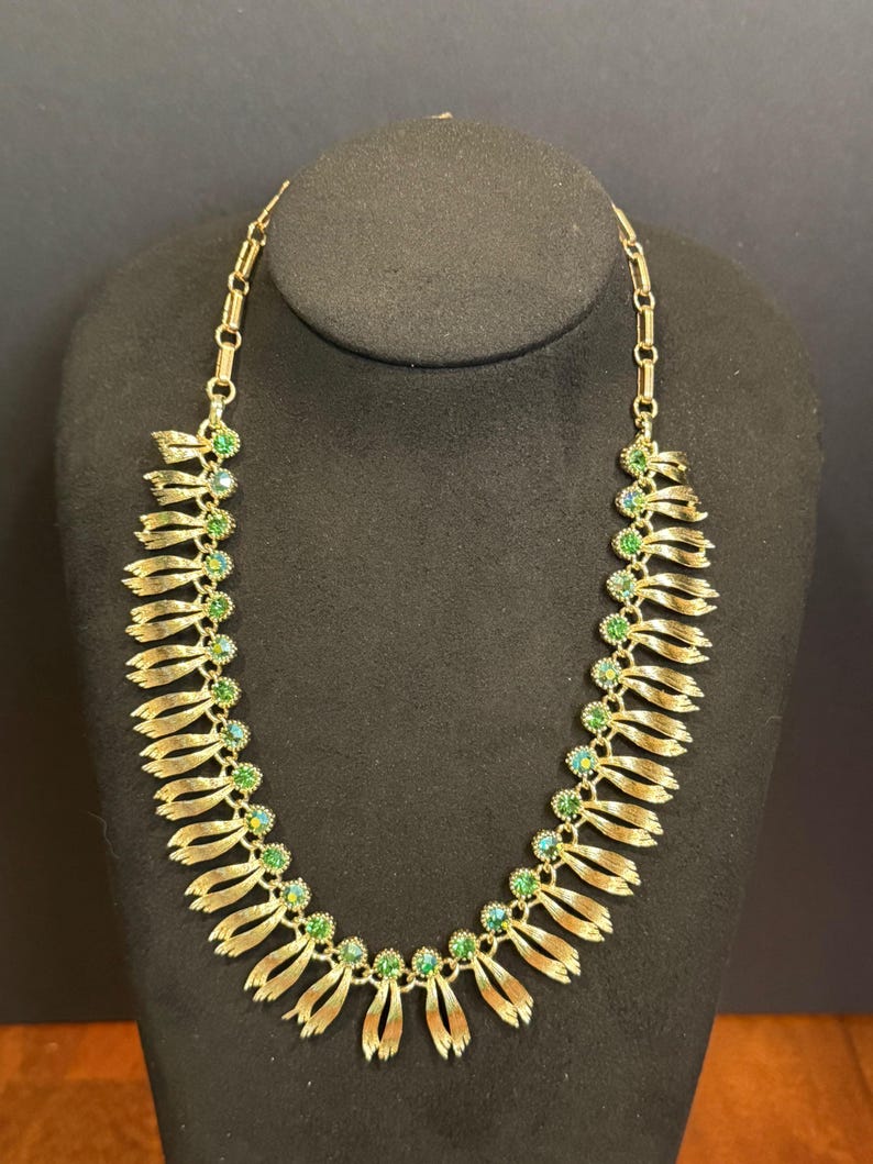 May include: Gold-toned necklace with a series of layered, ribbon-like elements. Each ribbon section is adorned with a cluster of green and teal-colored gemstones. The necklace is displayed on a black mannequin, showcasing its design and details.