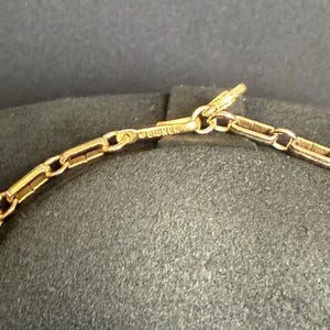 May include: Close-up of a gold-colored necklace chain with a rectangular link design. The word "LISNER" is stamped on the clasp. The chain is displayed against a dark background.