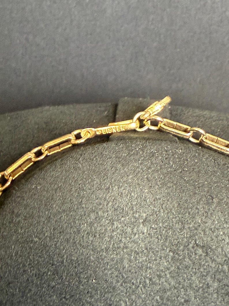 May include: Close-up of a gold-colored necklace chain with a rectangular link design. The word "LISNER" is stamped on the clasp. The chain is displayed against a dark background.