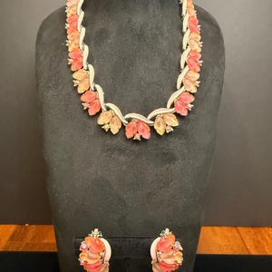 May include: A vintage jewelry set featuring a necklace and matching earrings. The necklace and earrings showcase a floral design with orange and peach-colored petals. The metal components are silver-toned, and the set is displayed on a black mannequin.