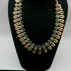 May include: A gold-toned necklace and matching earrings set. The necklace features a series of gold leaf-shaped pendants with green and blue crystal accents. The earrings are round, with a floral design and matching green and blue crystals.