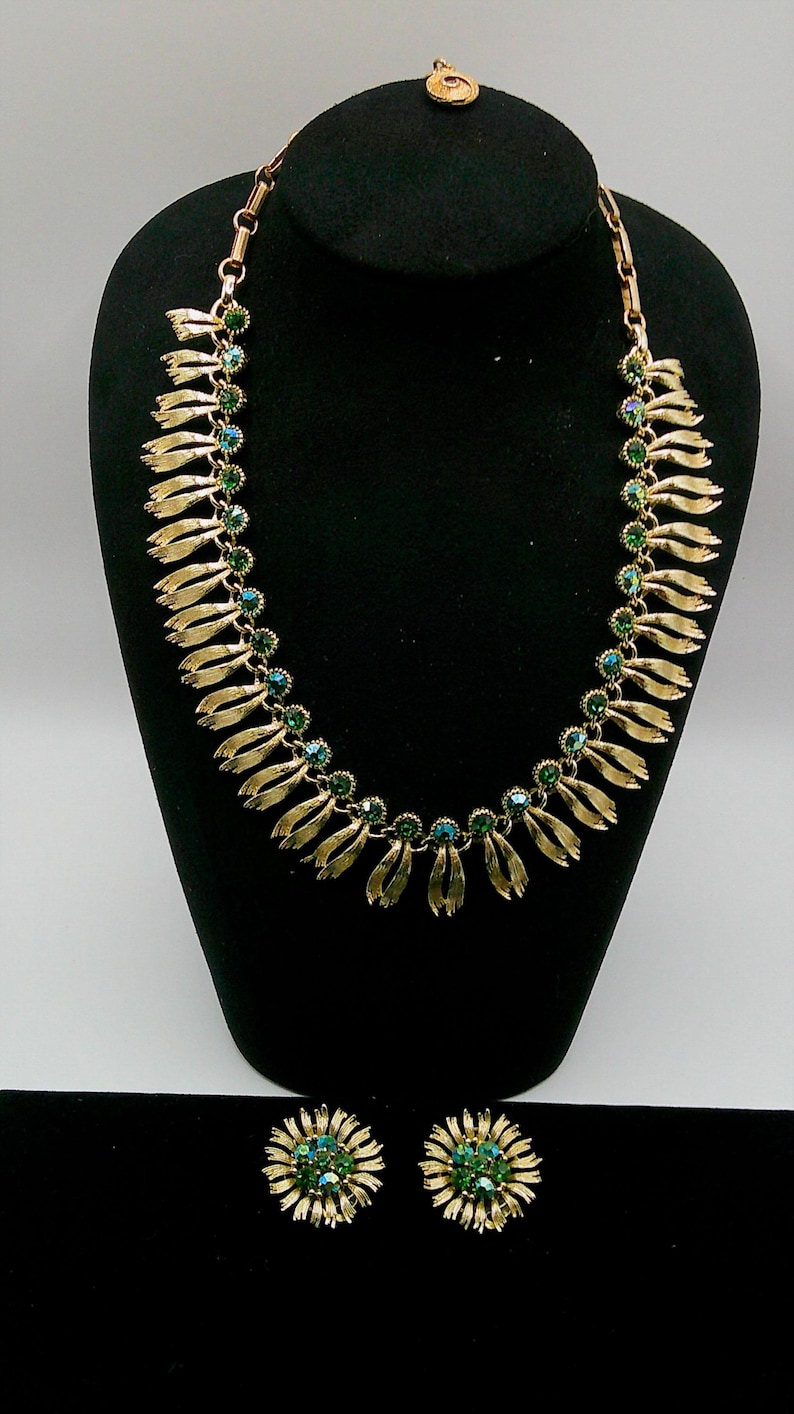 May include: A gold-toned necklace and matching earrings set. The necklace features a series of gold leaf-shaped pendants with green and blue crystal accents. The earrings are round, with a floral design and matching green and blue crystals.
