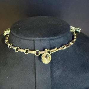 May include: A gold-toned necklace with a circular pendant and chain links. The necklace is displayed on a black mannequin. The pendant has a spiral design. The necklace has a clasp closure.