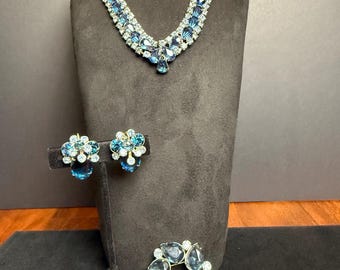 May include: A vintage jewelry set featuring a necklace, earrings, and a brooch. The necklace and earrings have blue teardrop-shaped stones and clear rhinestones. The brooch is a circular design with blue stones. The jewelry is displayed on a black mannequin.