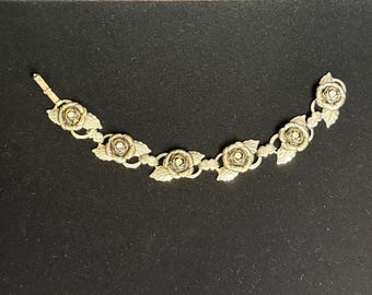 Peut inclure: Un bracelet vintage avec un motif floral. Le bracelet est composé d'éléments en forme de roses reliés entre eux, avec de petits accents ronds au centre de chaque fleur. Le métal est de couleur dorée et le bracelet est posé sur un fond sombre.