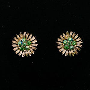 May include: A pair of gold-toned, flower-shaped earrings. Each earring features a cluster of green gemstones at the center, surrounded by radiating, textured gold petals. The earrings are set against a black background.